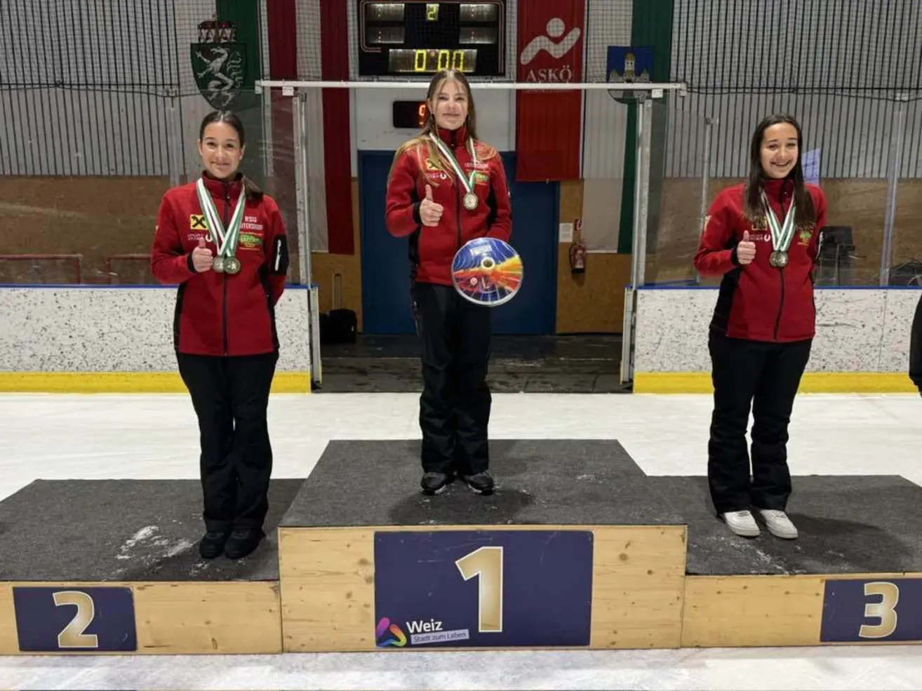 Three girls stand on a podium, holding medals, with one girl in the middle holding a round object. The setting is an ice rink.