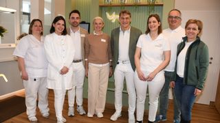 Eine Gruppe von Menschen in weißen Laborkittel und Anzügen, die zusammenstehen und für ein Foto in einem Raum mit Holzböden und grünen Wänden lächeln.