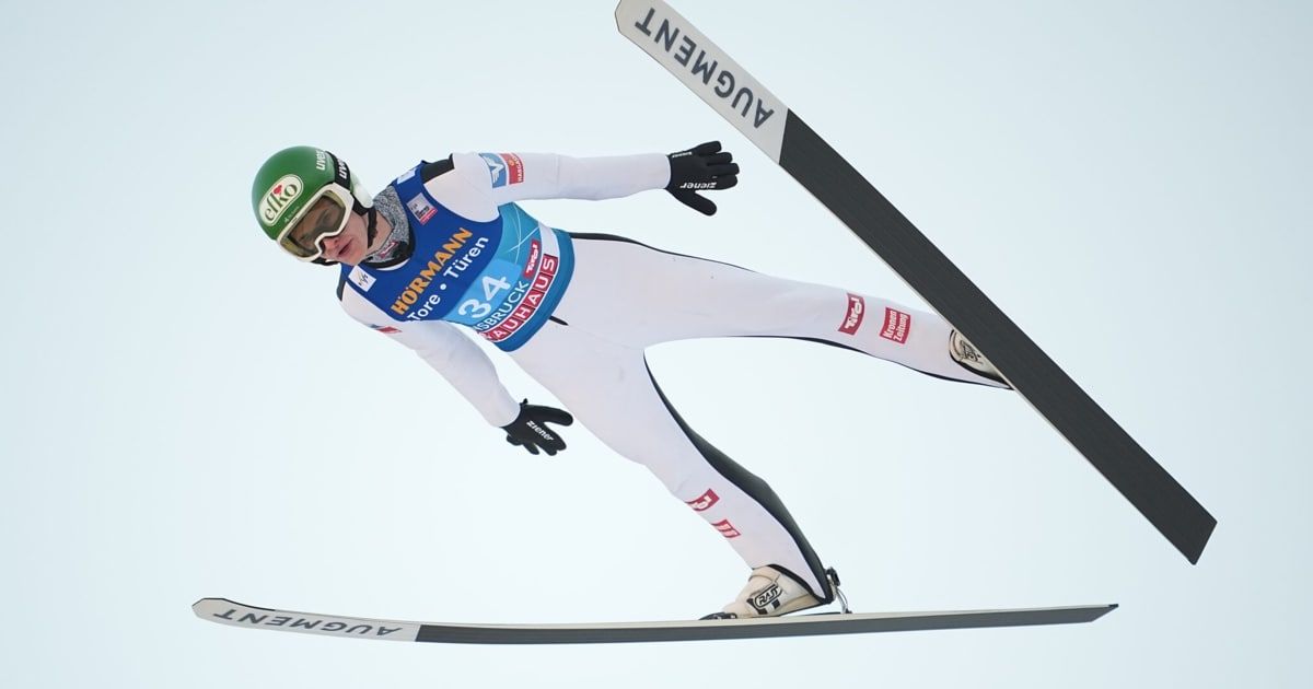 Ein Skispringer ist in der Luft mit weit gespreizten Skiern, trägt einen weißen und blauen Anzug und einen grünen Helm mit Schutzbrille.
