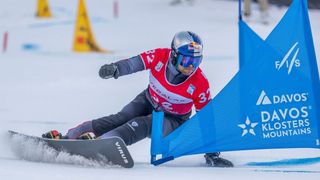 Ein Skifahrer in einem roten und grauen Outfit und Helm fährt den Schneehang hinunter, mit der Nummer 32 auf dem Rücken.