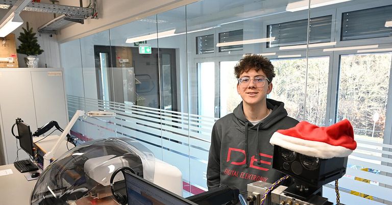 Ein Mann in einer grauen Kapuzenjacke mit 'DE' darauf gedruckt steht vor einer Glaswand und lächelt. Hinter ihm gibt es Reflexionen eines Raumes mit einem Fenster und einem Ausgangsschild. Auf der linken Seite eine Maschine mit einem Monitor und Kopfhörern. Auf der rechten Seite eine rote und weiße Weihnachtsmütze.