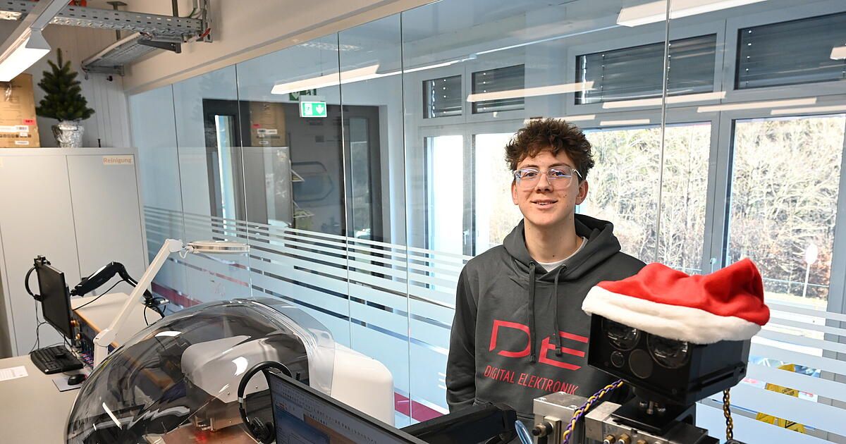 Ein Mann in einer grauen Kapuzenjacke mit 'DE' darauf gedruckt steht vor einer Glaswand und lächelt. Hinter ihm gibt es Reflexionen eines Raumes mit einem Fenster und einem Ausgangsschild. Auf der linken Seite eine Maschine mit einem Monitor und Kopfhörern. Auf der rechten Seite eine rote und weiße Weihnachtsmütze.