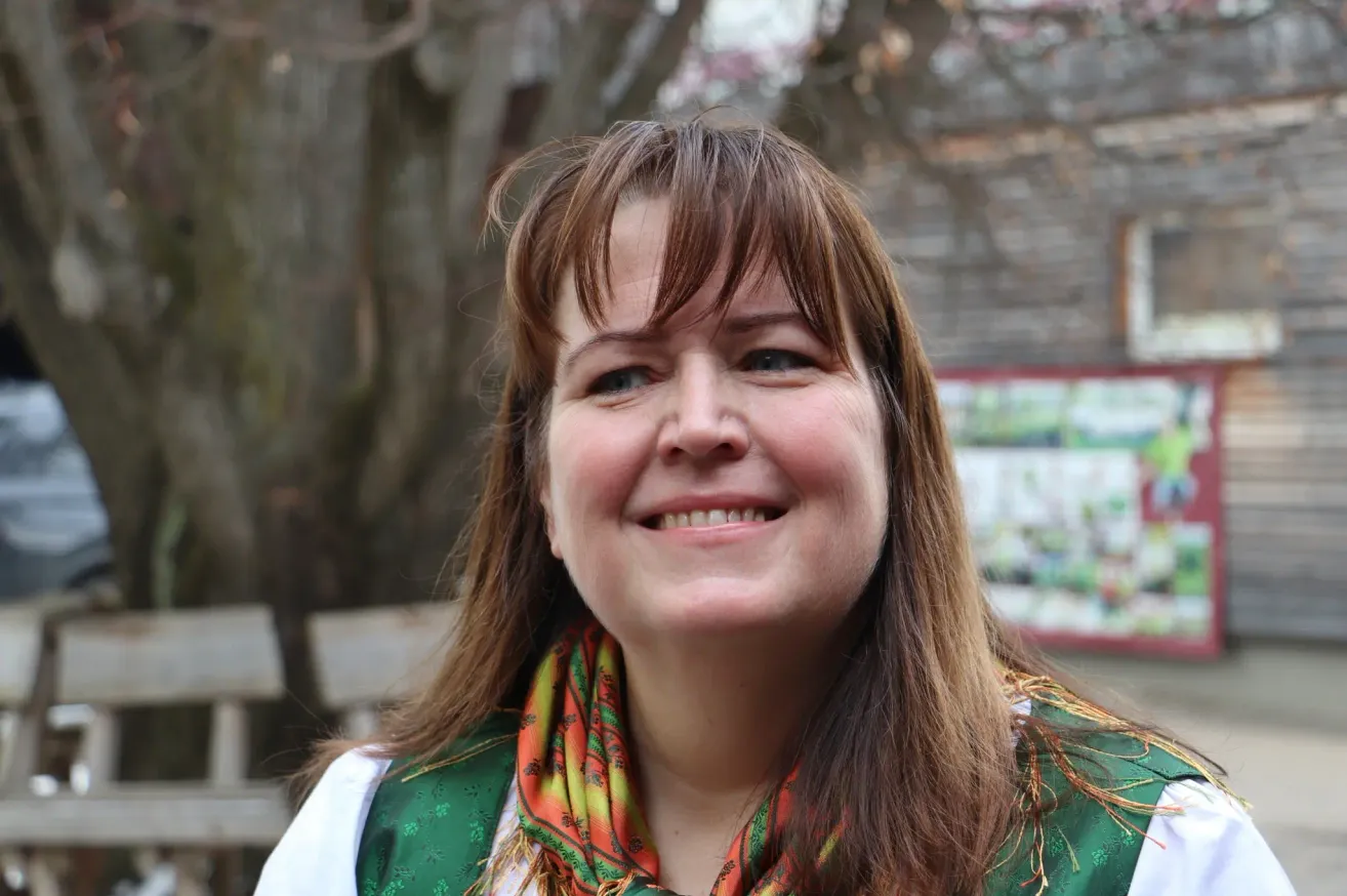 A woman with brown hair and a scarf smiles at the camera, standing in front of a wooden bench and trees.