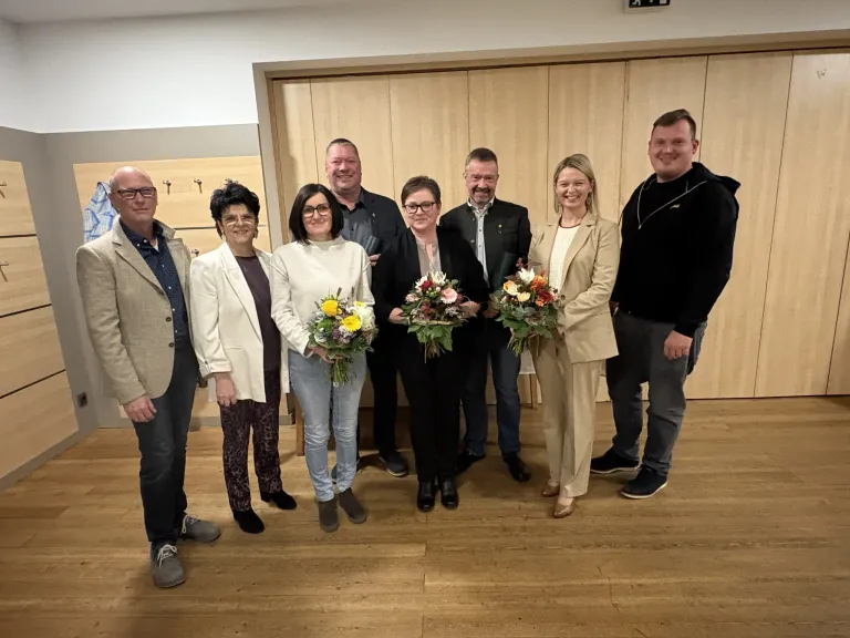 A group of seven individuals, all smiling, stand together indoors. They are holding flower bouquets, dressed in formal attire, and appear to be celebrating an event. Behind them is a wooden wall.