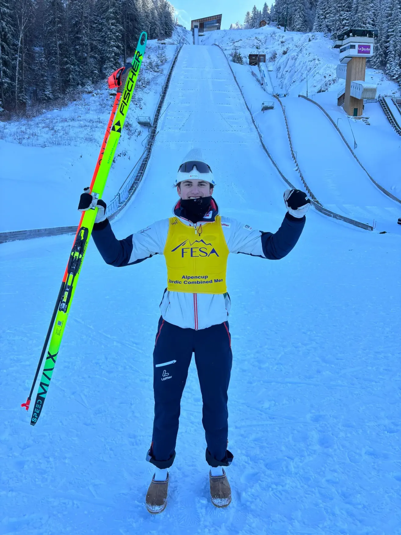 A man stands in the snow holding skis. He wears a yellow vest with the word FESA and a mask. Behind him, a ski jump is visible.