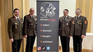 Drei Männer in Militäruniformen stehen neben einem großen Banner mit einem Logo, dem Text 'Feuerwehr Blindenmarkt' und der Phrase 'retten, löschen, bergen, schützen'. Das Banner ist dunkelgrau mit weißem und orangefarbenem Text und Symbolen.