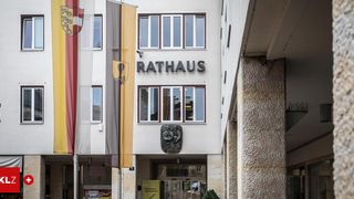Ein Gebäude mit dem Wort 'Rathaus' an der Wand, mit Fahnen auf der linken Seite, einer Skulptur unter dem Fenster und Glastüren am Eingang.