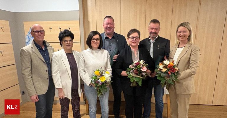 Eine Gruppe von Menschen, möglicherweise eine Familie, steht zusammen in einem Raum mit Holzwänden. Sie alle lächeln und halten Blumen. Sie sind in Freizeitkleidung gekleidet.
