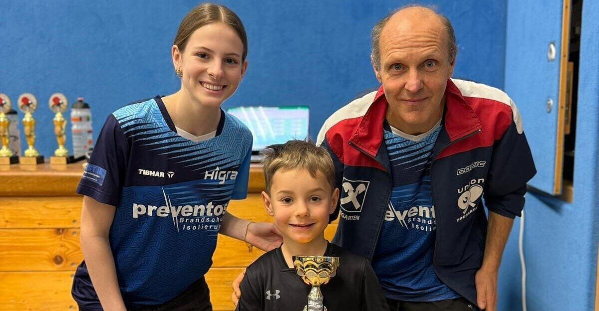 Three individuals pose for a photo. A young boy holds a trophy, with a smiling woman and an older man beside him. The woman wears a blue 'Prevento' jersey.