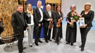 Six individuals are standing in front of a blue backdrop. They are holding violins. They are all smiling and appear to be posing for a photograph. Behind them is a white backdrop.