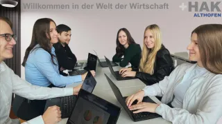 Mehrere Personen sitzen um einen Tisch herum, jede benutzt Laptops. Der Tisch ist voller Laptops und eine Tafel dahinter hat fremde Schrift.