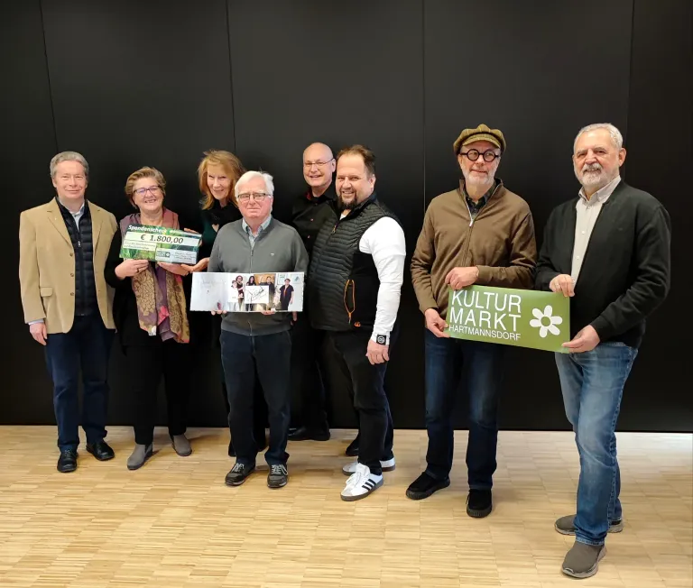 Sieben Personen stehen zusammen, lächeln und halten ein Plakat und ein Büchlein. Sie scheinen ein Ereignis zu feiern.
