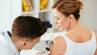 Ein Mann in einem Laborkittel untersucht die Schulter einer Frau mit einer Lupe. Sie trägt ein weisses ärmelloses Oberteil.