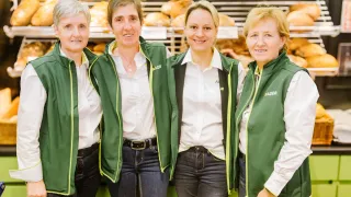 Vier Frauen, gekleidet in grünen Westen und weißen Hemden, lächeln und posieren für ein Foto vor einer Bäckerei-Auslage.