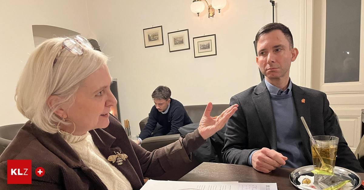 Eine Frau und ein Mann sitzen an einem Tisch und unterhalten sich. Ein anderer Mann sitzt hinter ihnen auf einer Couch. Darüber hängen drei gerahmte Bilder an der Wand.