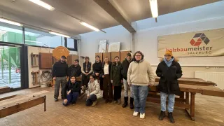Eine Gruppe von Menschen posiert für ein Foto in einer Werkstatt. Sie stehen und sitzen auf Holzbänken. Ein Banner mit einem Logo ist an der Wand angebracht.