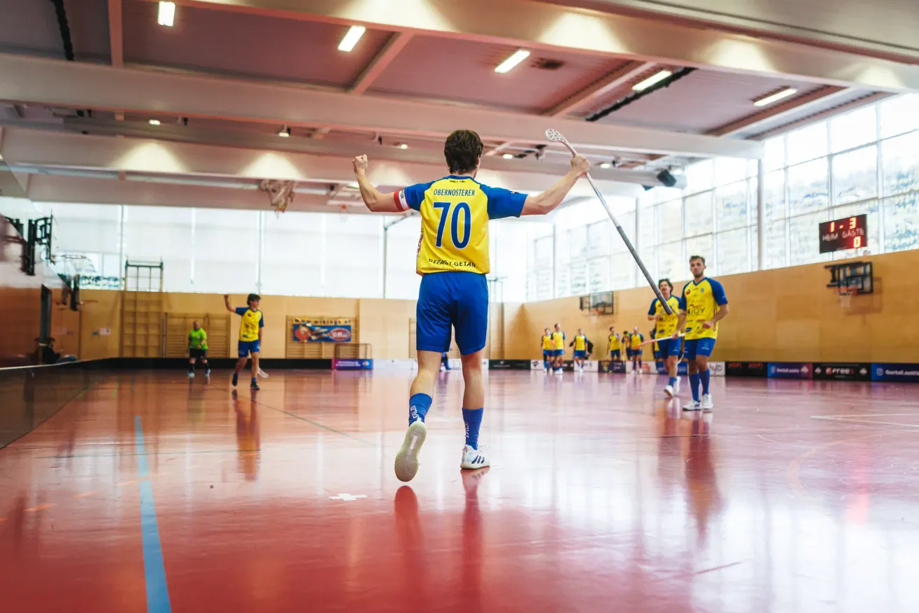 Ein Spieler in gelber und blauer Uniform feiert ein Tor bei einem Handballspiel mit Teamkollegen auf einem roten Spielfeld.