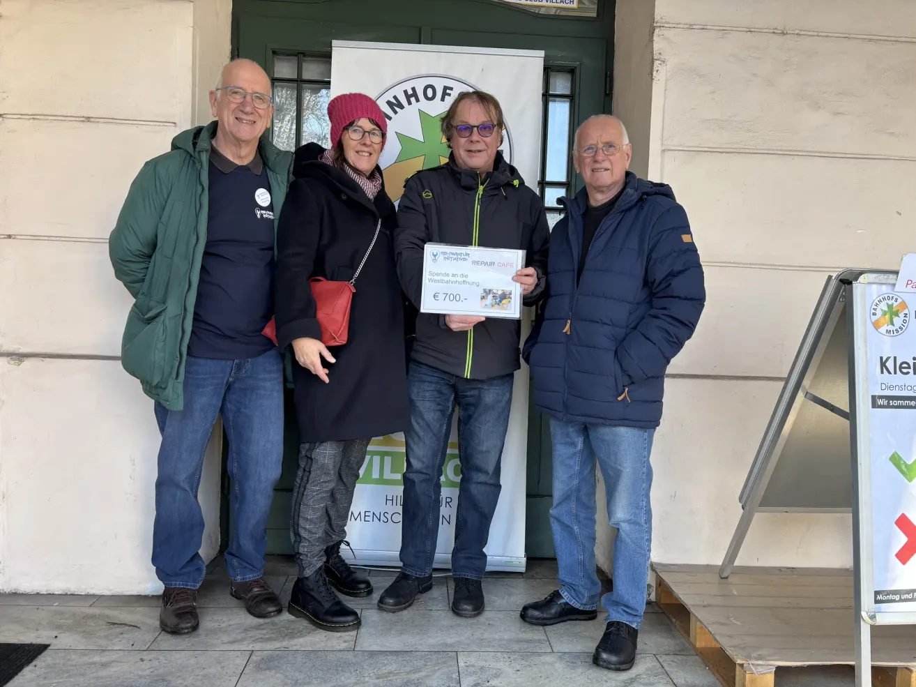 Vier Menschen posieren lächelnd für ein Foto mit einem Scheck in der Mitte. Der Scheck ist mit 'Spenden an die Werkstatthof' und einem Betrag von 700 Euro beschriftet. Sie stehen vor einem Gebäude mit einem Banner.