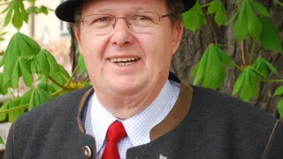 Ein Mann mit Hut, Brille und roter Krawatte lächelt mit einer Blume auf der Brust. Er steht draußen in der Nähe eines Baumes.