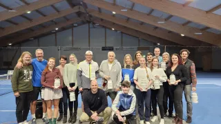 Eine Gruppe von Menschen steht in einer Turnhalle mit einer Holzdecke. Sie halten alle weiße Behälter und lächeln. Einige von ihnen tragen Turnschuhe und Jacken. Der Boden ist grau. Die Decke hat viele Lichter. Es gibt Werbebanner an der Wand.