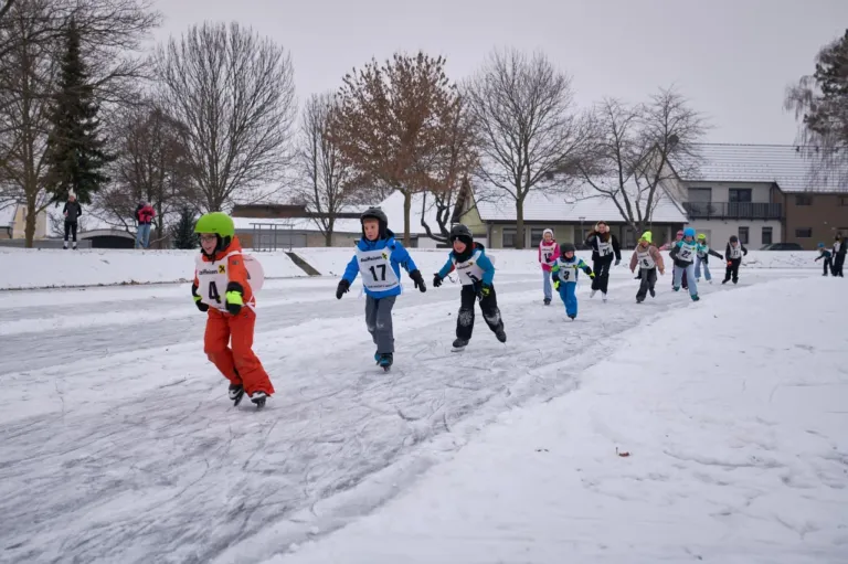 Kinder in Winterkleidung nehmen an einem Schneerennen teil. Ein Kind trägt einen grünen Helm und die Nummer 4, ein anderes trägt die Nummer 17. Bäume ohne Blätter und eine verschneite Landschaft sind im Hintergrund zu sehen.