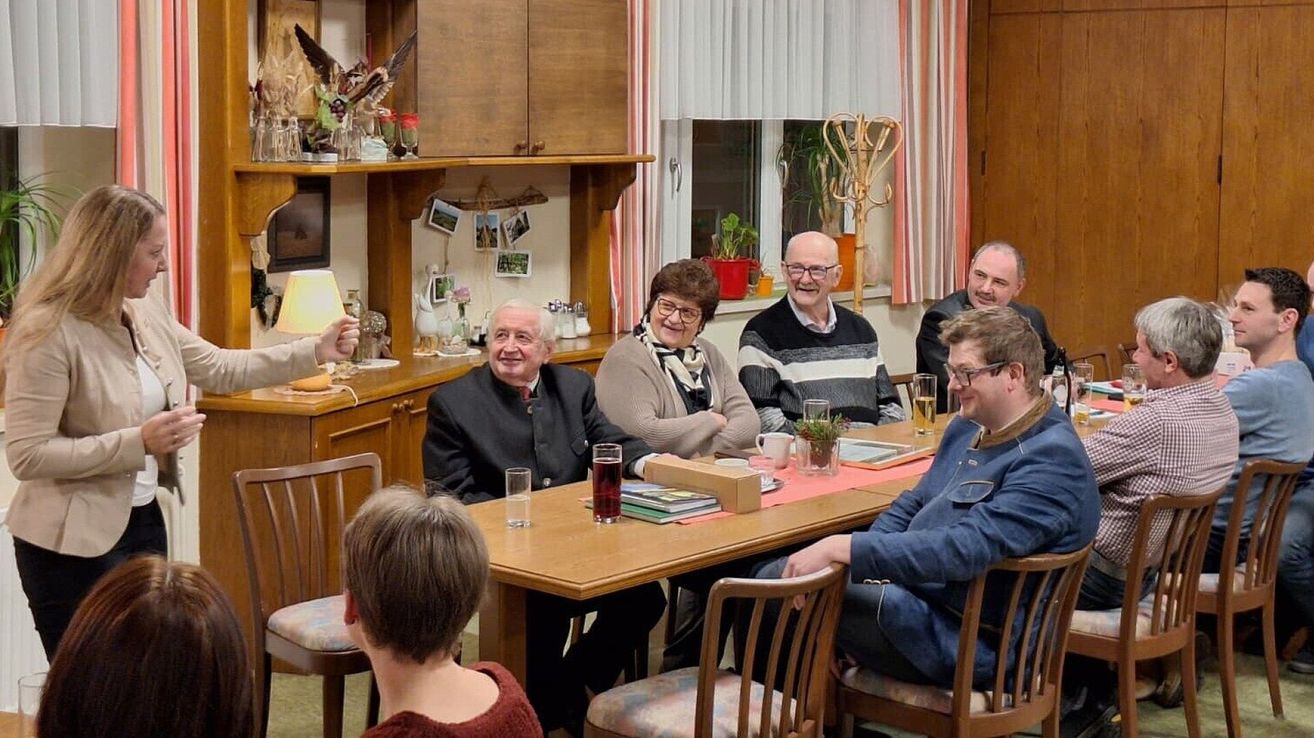 Eine Gruppe älterer Menschen sitzt lächelnd und plaudernd um einen Tisch. Auf dem Tisch stehen Gläser, Bücher und eine Lampe. Dahinter befindet sich eine Küche mit Schränken, einem Fenster mit Vorhängen und Topfpflanzen.