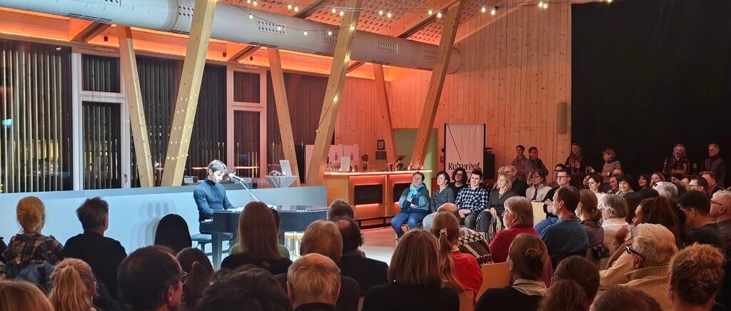A pianist plays on a grand piano at a cultural event, with a large audience seated in front of him. The room has wooden walls and ceiling with decorative lights. There is a banner with text and a wooden counter with a person standing behind it.