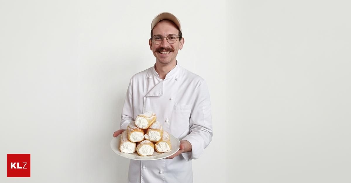 Ein Mann in einer weißen Kochuniform lächelt und hält einen Teller mit Sahne gefüllten Gebäckstücken. Er steht vor einem schlichten weißen Hintergrund.
