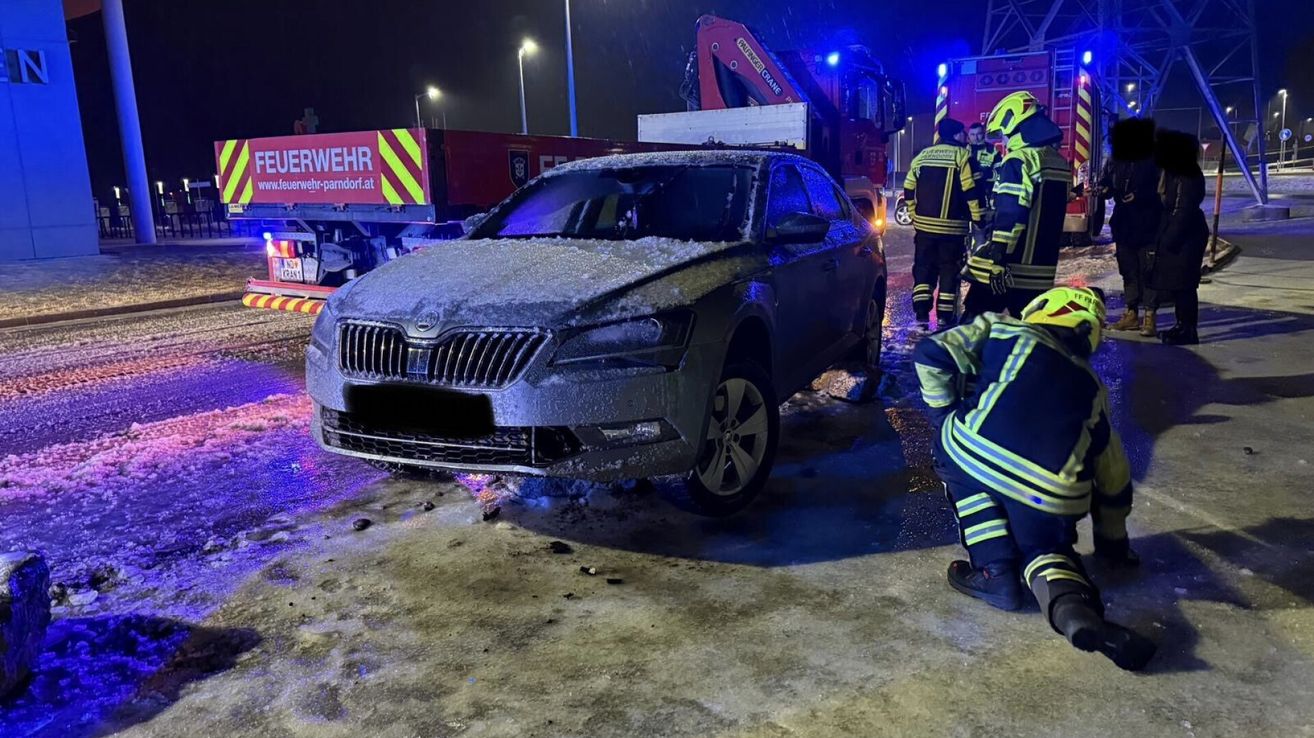 A damaged Skoda car covered in ice sits on a wet street at night. Emergency workers in yellow and green uniforms stand nearby.