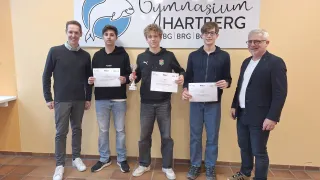 Fünf Personen, darunter drei mit Urkunden und einer mit einer Trophäe, posieren für ein Foto vor einem Gymnasium Hartberg-Schild.
