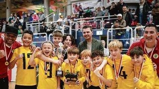 A group of young boys in yellow jerseys with numbers pose with a trophy and medals, smiling for the camera. A man in a green suit stands behind them. Spectators are seated in the background, watching.