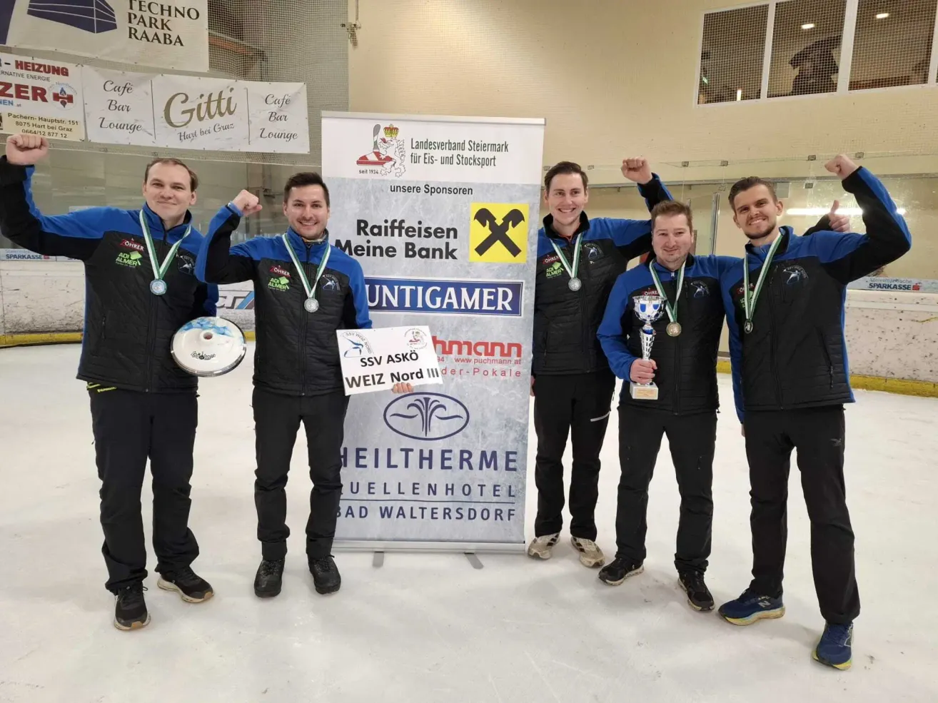 Fünf Männer stehen auf einer Eisbahn, tragen Medaillen und halten Pokale. Hinter ihnen zeigt ein Banner Sponsoren und ein Logo. Die Umgebung umfasst Banner für Cafés und Lounges.