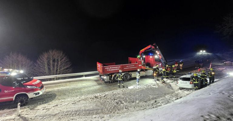Emergency personnel respond to a vehicle accident on a snowy road at night. A red truck with flashing lights is parked, and several people are attending to the scene.