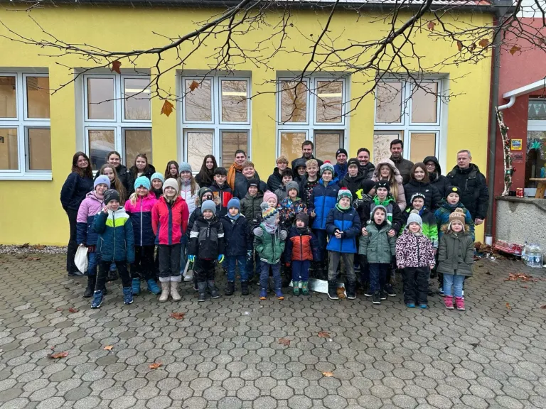A group of children and adults, dressed in winter clothing, poses for a photo in front of a yellow building with large windows.