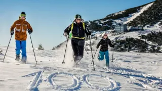 Two skiers on a snowy mountain, one wearing a green cap and sunglasses, the other in a checkered jacket. Behind them, a building with many windows.