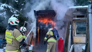 Firefighters are working to extinguish a fire in a wooden shed, with smoke and flames visible. One firefighter uses a hose while another has a fire extinguisher. Nearby, a red fire extinguisher is placed, and a helmet rests on a bucket.