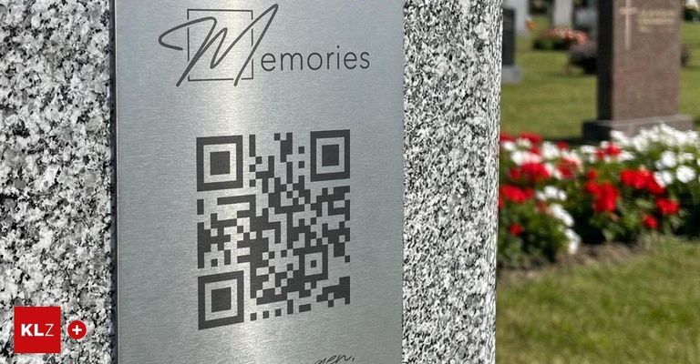 A metal plaque with the logo 'N Memories' and a QR code is mounted on a granite tombstone. Behind the tombstone, there are flowers and grass.