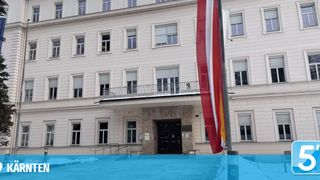 Ein weißes Gebäude mit vielen Fenstern und einer Flagge davor. Über dem Eingang befindet sich ein Schild.