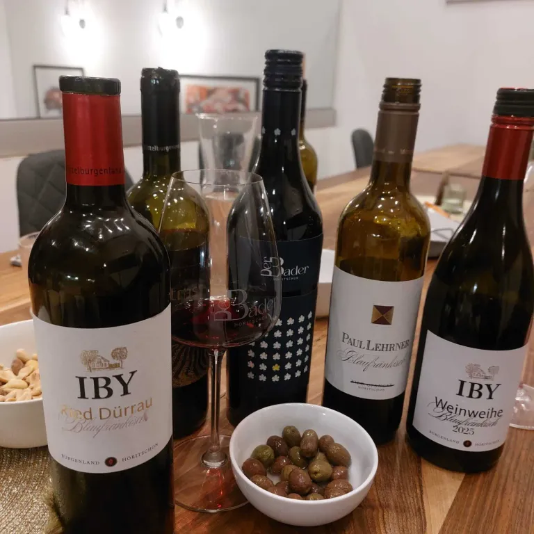 A wooden table displays several wine bottles, including IBY Red Durrau and Paul Lehrer. A wine glass filled with red wine stands between them. A bowl of olives is on the table, with a glass bowl of nuts in the background.