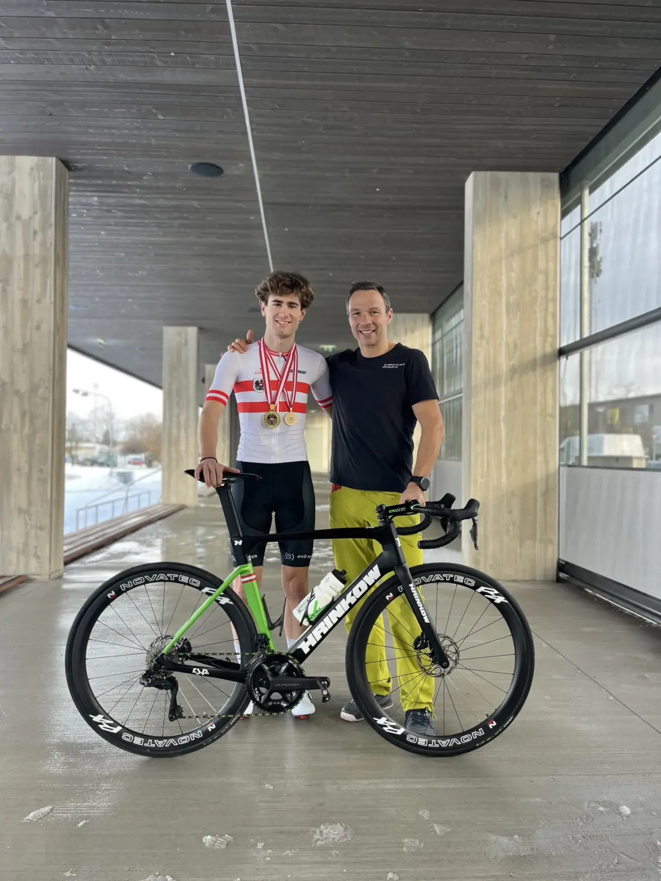 Zwei Männer stehen neben einem Hinkly-Fahrrad, einer mit einer Medaille um den Hals und der andere in einem schwarzen T-Shirt. Sie lächeln und scheinen für ein Foto zu posieren.