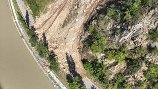 An aerial view shows a rocky hill with vegetation, a road on the left, and a river at the bottom.