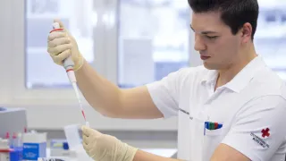 A man in a lab coat, wearing gloves, is holding a pipette and appears to be conducting an experiment in a laboratory.