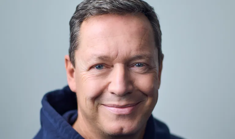 A man with graying hair, blue eyes, and a warm smile wearing a blue hoodie against a light background.