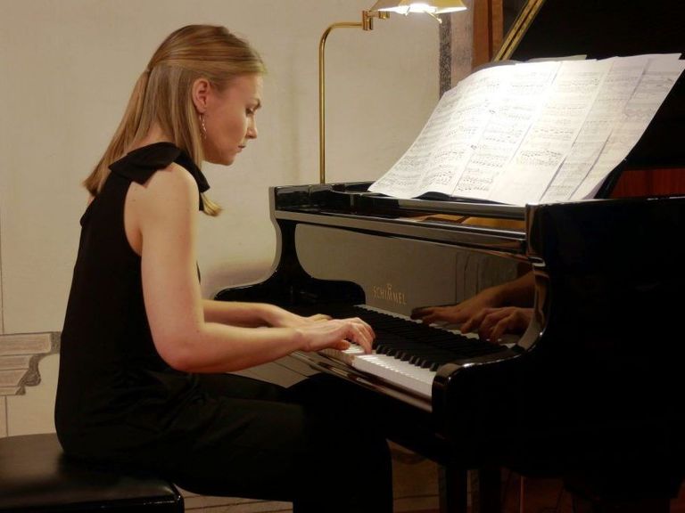 A woman is seated at a black grand piano, her hands on the keys. A music sheet is open on the piano. A lamp is mounted on the wall behind her.
