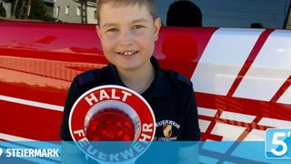 A young boy in a black uniform with red trim holds a red light, smiling and standing in front of a fire truck.