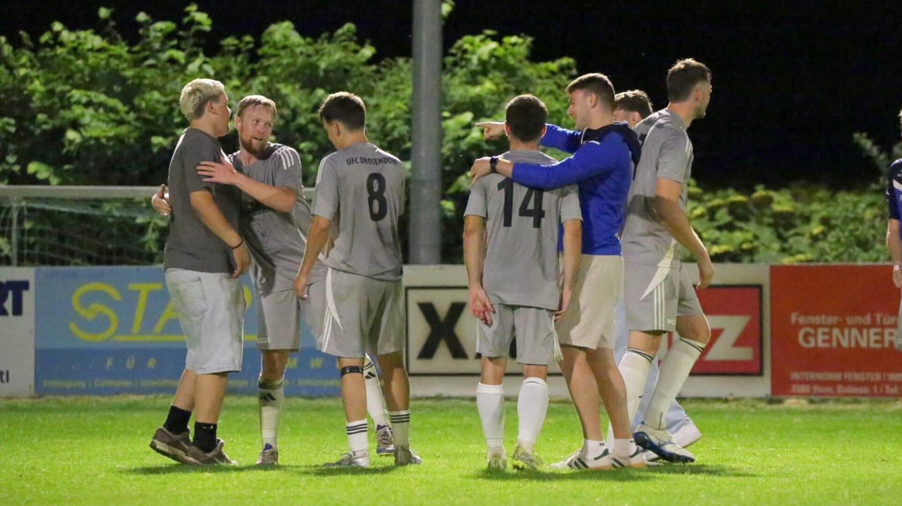Eine Gruppe von Fußballspielern in grauen Trikots versammelt sich auf dem Spielfeld, einer von ihnen zeigt, während die anderen aufmerksam zuhören.