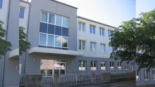 A modern building with glass windows and a sign reading 'Bundeshandelsakademie' and 'Bundeshandelsschule'. The entrance has double glass doors and a metal railing.