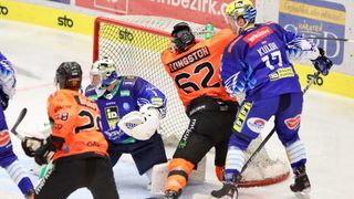 Ein Eishockeyspiel mit Spielern in Trikots mit den Nummern 28, 1, 62 und 77. Ein Spieler schießt, während ein anderer verteidigt. Der Torwart steht vor dem Netz.