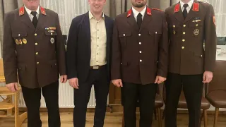 Four men stand together in formal attire. Three wear military uniforms, and one wears a business suit. They are standing in a room with wooden floors and white curtains.