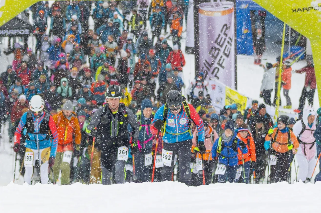 A group of skiers with bib numbers run through snow, with spectators in the background. Some wear helmets and carry ski poles. There's a banner with text in the snow.